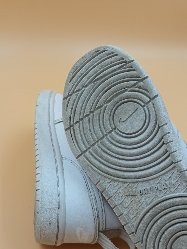 以下が含まれることがあります： 白い革のハイカットスニーカーで、白いゴム製のソールが付いています。ソールには円形の模様があり、底には「All Day Play」という文字が印刷されています。