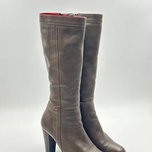 Könnte beinhalten: Braune Kniehochstiefel aus Leder mit Blockabsatz. Die Stiefel haben eine runde Spitze, einen Reißverschluss an der Innenseite und ein rotes Futter. Sie sind aus glattem Leder gefertigt und haben eine Plateausohle. Absatzhöhe ca. 8 cm.