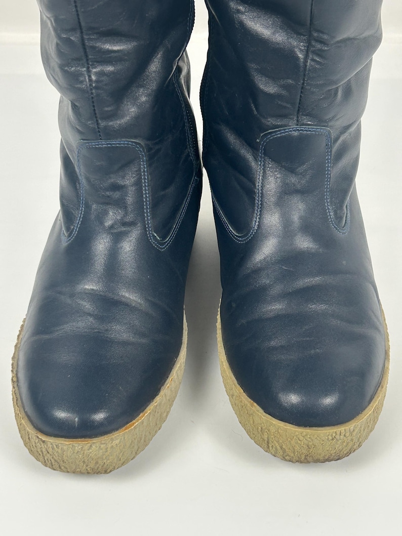 May include: A pair of navy blue, knee-high boots. The boots have a rounded toe and a thick, light brown sole. The upper portion of the boots is made of a smooth, slightly textured material.