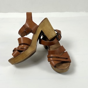 May include: A pair of brown leather platform sandals with wooden block heels. The sandals feature multiple straps across the foot and ankle straps with buckles. The heels are approximately 4 inches high.