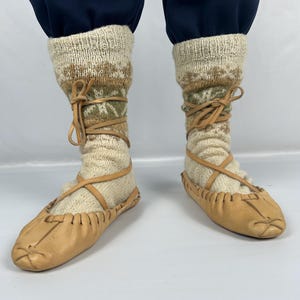 Puede incluir: Un par de zapatos tradicionales de cuero con calcetines de punto color crema. Los calcetines presentan un estampado marrón y verde y están atados con correas de cuero. Los zapatos son de color marrón claro y tienen un diseño cosido.