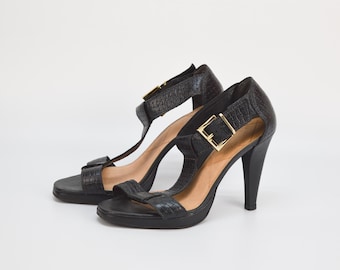 May include: A pair of black leather heeled sandals with gold buckles. The sandals have a t-strap design and an open toe.