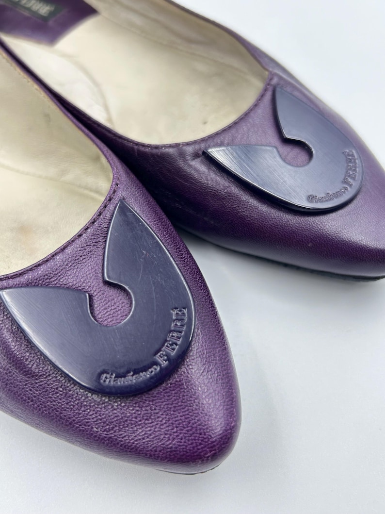 Op de afbeelding: Een paar paarse leren ballerina's met een donkerpaarse decoratieve accent op de neus. Het accent heeft een unieke vorm en de woorden "Gianfranco Ferre" staan erop gedrukt. De schoenen staan op een witte achtergrond.