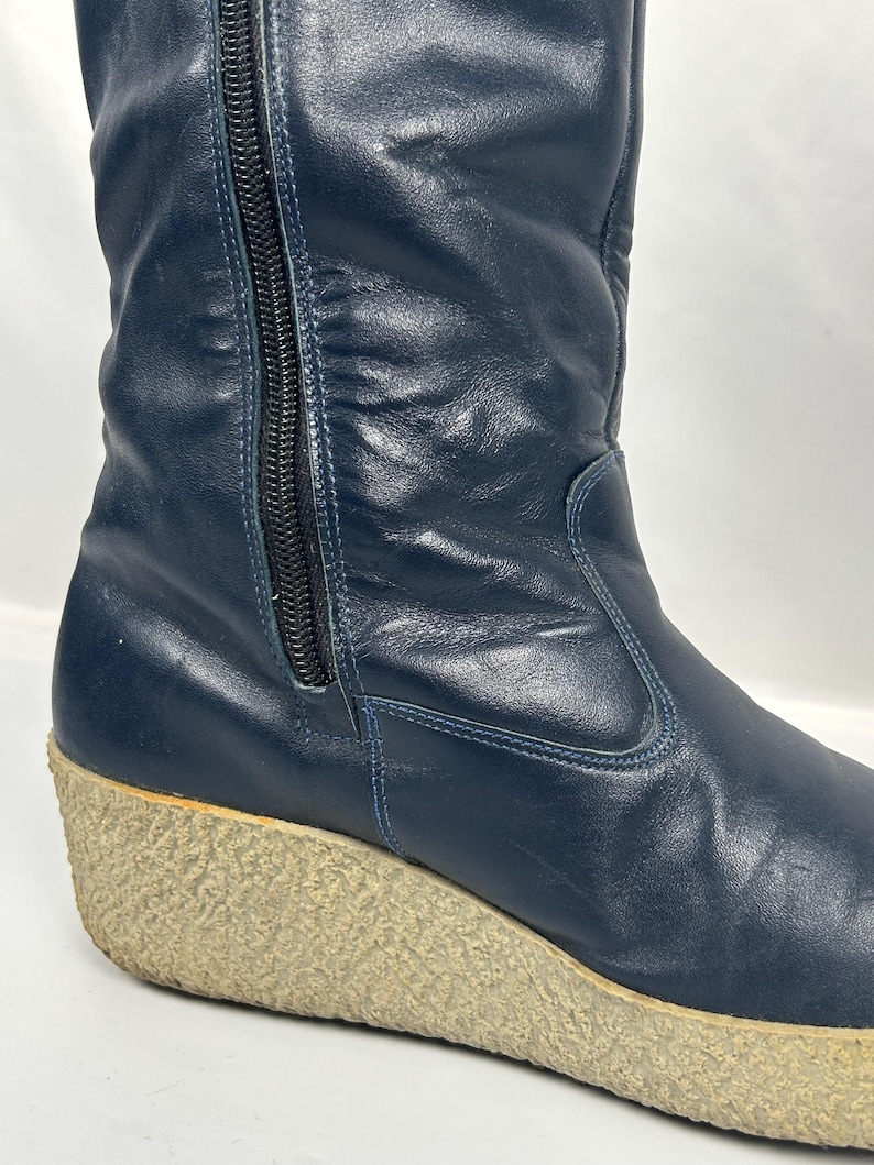 May include: Navy blue leather boot with a wedge sole. The boot features a side zipper and a textured, light-colored sole. The leather has a slight sheen, and the boot appears to be vintage.