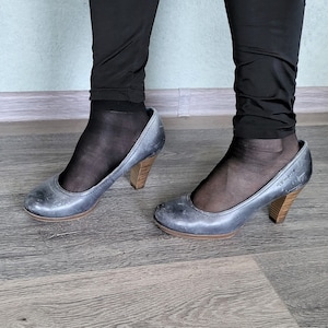Könnte beinhalten: Ein Paar graue Lederschuhe mit Absatz und Holzabsätzen. Die Schuhe werden mit schwarzen Strumpfhosen getragen.