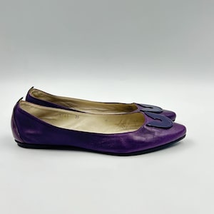 Op de afbeelding: Een paar paarse leren ballerina's met een donkerblauwe decoratieve accent op de neus. De schoenen zijn maat 36. De ballerina's staan op een witte achtergrond.