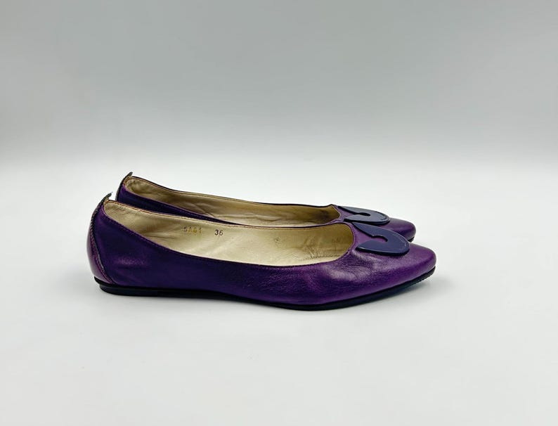 Op de afbeelding: Een paar paarse leren ballerina's met een donkerblauwe decoratieve accent op de neus. De schoenen zijn maat 36. De ballerina's staan op een witte achtergrond.