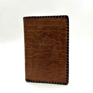 May include: A brown leather wallet with a detailed embossed design of a building facade. The wallet has black stitching along the edges and a rectangular shape. The design appears to be a historical landmark.