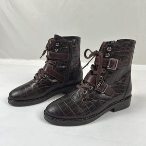 May include: A pair of dark brown ankle boots featuring a crocodile-embossed texture, lace-up fronts with matching brown laces, and two decorative straps across the upper shaft secured with silver-toned metal buckles and side zippers.