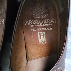 May include: Close-up of the inside of a brown leather shoe with the text "ARISTOKRAT den svenska herrskon med l&auml;derbindsulja" and a logo of a stylized "H" with the word "vidd" below it.