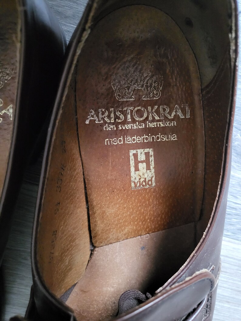 May include: Close-up of the inside of a brown leather shoe with the text "ARISTOKRAT den svenska herrskon med l&auml;derbindsulja" and a logo of a stylized "H" with the word "vidd" below it.
