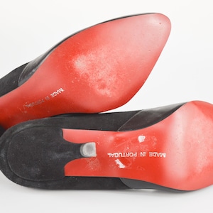 May include: Close-up of the red soles of a pair of black suede heeled shoes. The shoes are labeled "Made in Portugal".