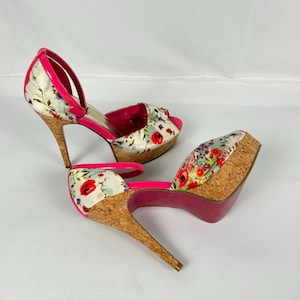 Puede incluir: Sandalias de tacón alto con estampado floral. Los zapatos tienen plataforma y tacón de corcho, con ribete y correas de color rosa brillante. El estampado floral incluye amapolas rojas y hojas verdes.