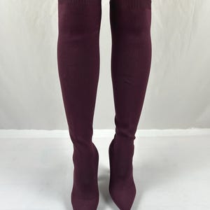 May include: A pair of dark burgundy over-the-knee boots crafted from a stretch knit fabric. These boots feature a pointed toe design and a ribbed knit cuff at the top edge, extending well above the knee with a form-fitting silhouette.
