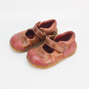 以下が含まれることがあります： 赤色のアクセントと装飾的なステッチが施された、茶色の革製の子供用靴のペア。靴にはバックル留めのストラップが付いています。
