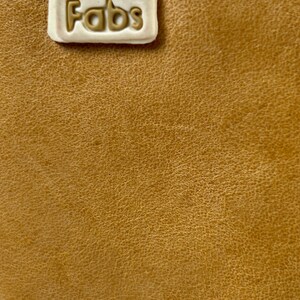 Puede incluir: Primer plano de una superficie de cuero bronceado texturizado. Una etiqueta rectangular blanquecina est&aacute; cosida en la parte superior, con la palabra "Fabs" en relieve en un color bronceado a juego. El cuero tiene una veta fina y natural.