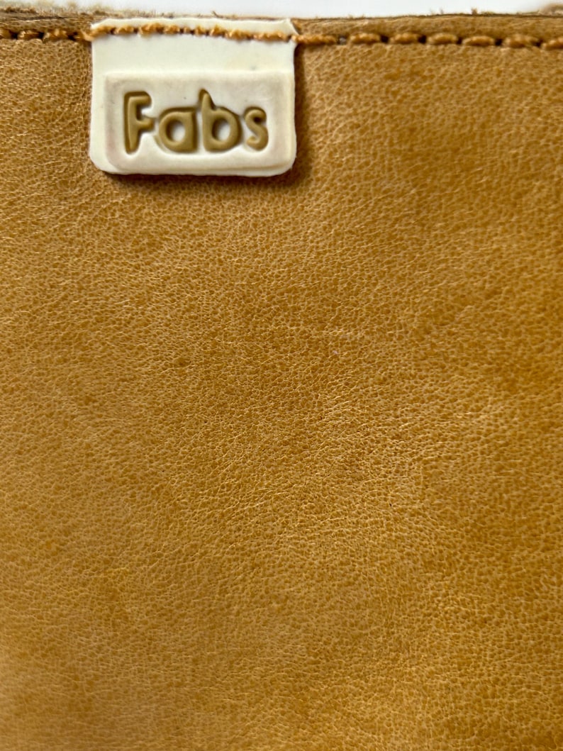 Puede incluir: Primer plano de una superficie de cuero bronceado texturizado. Una etiqueta rectangular blanquecina est&aacute; cosida en la parte superior, con la palabra "Fabs" en relieve en un color bronceado a juego. El cuero tiene una veta fina y natural.