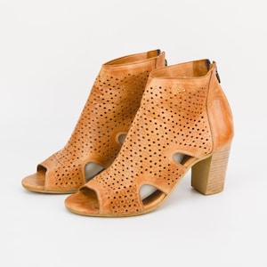 Könnte beinhalten: Ein Paar braune Ledersandalen mit Absatz, Ausschnitten und einem perforierten Design. Die Sandalen haben einen Blockabsatz und eine offene Zehenpartie.