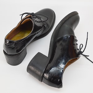 Puede incluir: Un par de zapatos de tac&oacute;n de charol negro con cordones. Los zapatos tienen un tac&oacute;n grueso y una punta redonda.