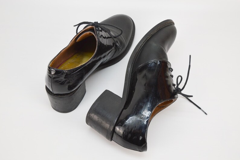 Puede incluir: Un par de zapatos de tac&oacute;n de charol negro con cordones. Los zapatos tienen un tac&oacute;n grueso y una punta redonda.