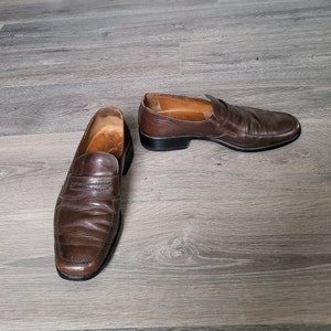 May include: A pair of brown leather loafers with a slip-on design. The loafers have a slightly worn look and are laid out on a wooden floor.