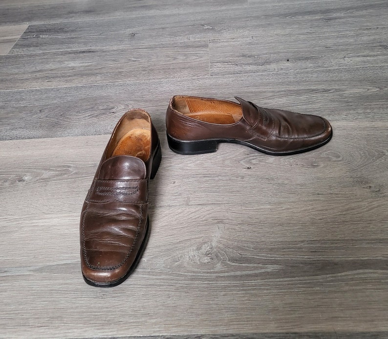 May include: A pair of brown leather loafers with a slip-on design. The loafers have a slightly worn look and are laid out on a wooden floor.