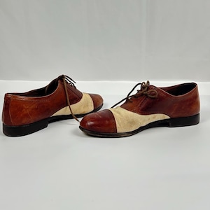 May include: A pair of two-tone brown and beige oxford shoes with laces. The shoes have a classic design with a leather upper and a contrasting beige fabric toe. The soles are black.