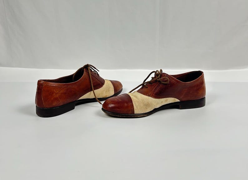 May include: A pair of two-tone brown and beige oxford shoes with laces. The shoes have a classic design with a leather upper and a contrasting beige fabric toe. The soles are black.
