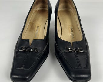 May include: A pair of black leather heeled shoes with square toes. Each shoe has a silver-colored buckle detail on the vamp. The brand name "SIVELGA" is visible inside the shoe.