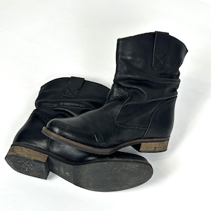 Puede incluir: Un par de botines de cuero negro con un aspecto desgastado y envejecido. Los botines tienen un tacón bajo y un diseño ligeramente arrugado alrededor del tobillo. Las suelas muestran signos de desgaste.