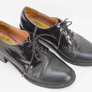 Puede incluir: Un par de zapatos de tac&oacute;n de cuero negro con cordones. Un zapato tiene un acabado de charol.