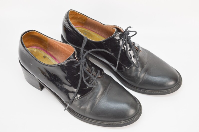 Puede incluir: Un par de zapatos de tac&oacute;n de cuero negro con cordones. Un zapato tiene un acabado de charol.