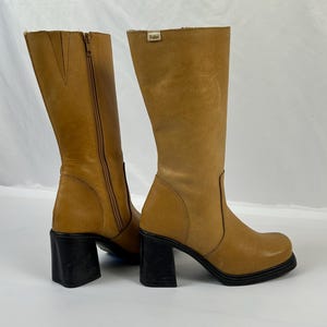 Puede incluir: Botas altas de cuero color canela con tacones de bloque negros. Una bota tiene una cremallera lateral. Las botas tienen punta cuadrada y una peque&ntilde;a etiqueta en la parte superior. Los tacones miden unos 7,6 cm.