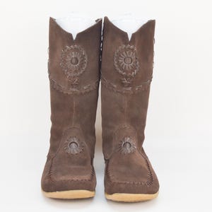 May include: A pair of brown suede boots with decorative floral cutouts. The boots have a mid-calf height, a rounded toe, and a stitched sole. The boots have a soft, textured appearance and a neutral color palette.