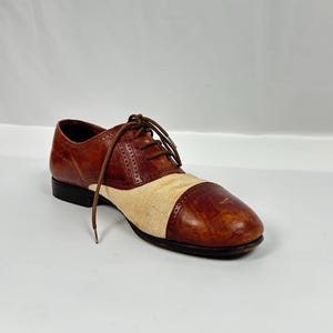 May include: A single brown and beige oxford shoe with a cap toe. The shoe features a leather upper with a beige fabric panel and brown laces. The shoe has a black sole and heel.