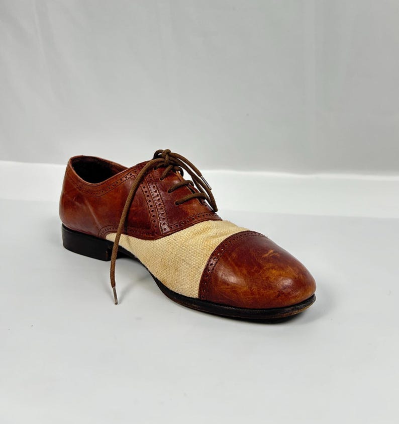 May include: A single brown and beige oxford shoe with a cap toe. The shoe features a leather upper with a beige fabric panel and brown laces. The shoe has a black sole and heel.