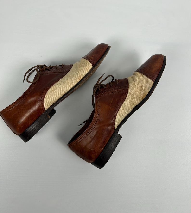 May include: A pair of two-tone oxford shoes. The shoes feature brown leather uppers with tan fabric panels on the sides and toes. The shoes have brown laces and black soles.