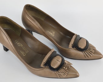 May include: A pair of brown leather heeled shoes with a pointed toe. The shoes feature a black metal buckle with a fringe detail. The shoes are labeled "Salvatore Ferragamo" on the insole.