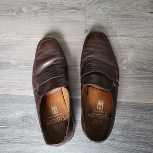 May include: A pair of brown leather loafers with a stitched design. The loafers have a leather insole with the text "Aristocrat" and "Made in Italy" printed on them.