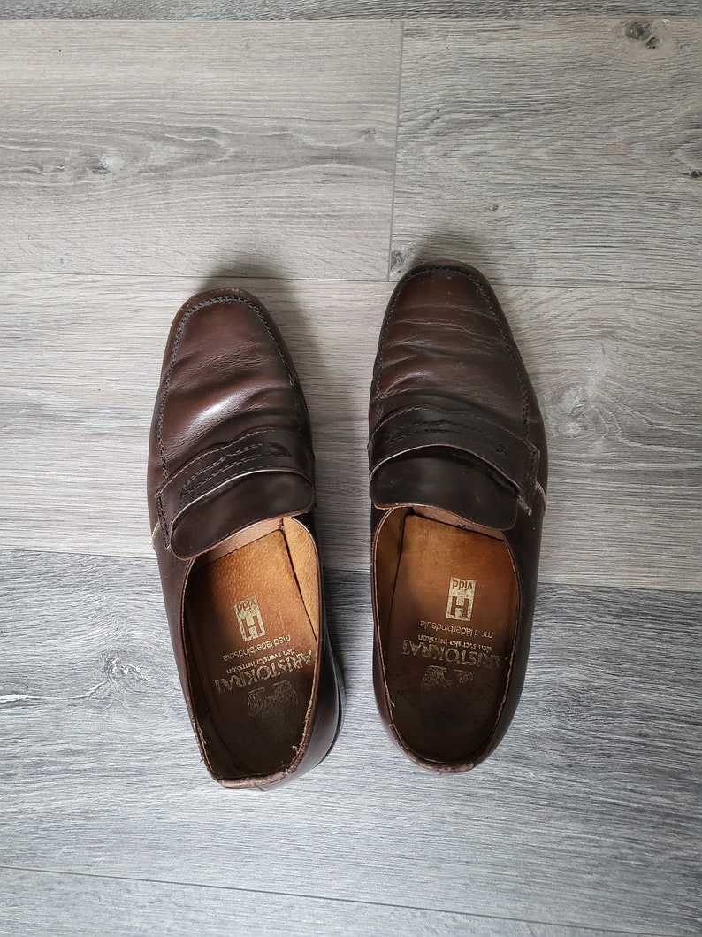 May include: A pair of brown leather loafers with a stitched design. The loafers have a leather insole with the text "Aristocrat" and "Made in Italy" printed on them.