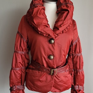 Puede incluir: Una chaqueta acolchada roja con un cuello grande y fruncido, un cinturón y botones decorativos. La chaqueta tiene un diseño acolchado en las mangas y una silueta ajustada.