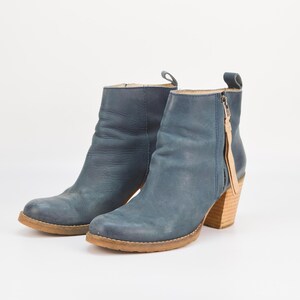 May include: A pair of blue leather ankle boots with a stacked wooden heel. The boots have a side zipper with a tan leather pull and a round toe. The soles are a light brown color.