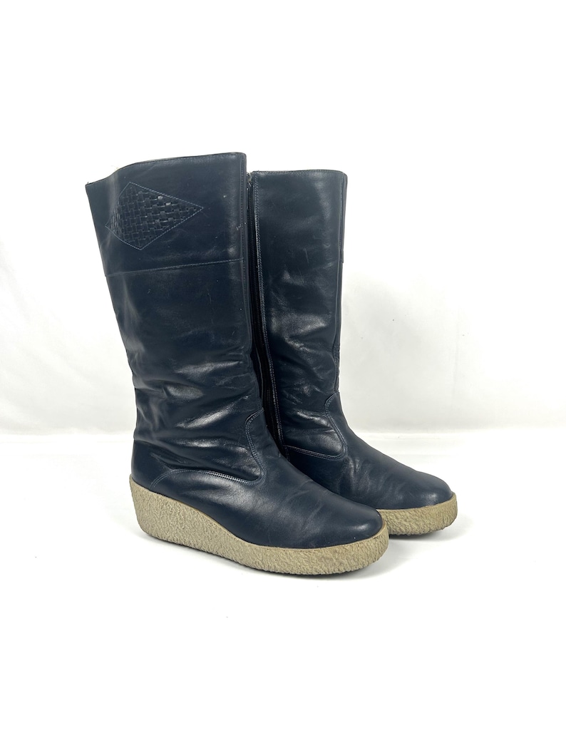 May include: A pair of tall, black leather boots with a wedge sole. The boots have a side zipper and a decorative diamond-shaped pattern near the top. The soles are a light tan color.