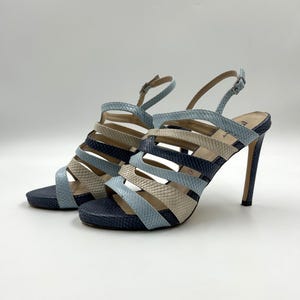 May include: A pair of high-heeled sandals with a strappy design. The sandals feature a combination of light blue, navy, and beige faux snakeskin. The heels are approximately 4 inches tall.