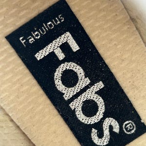 Puede incluir: Primer plano de una etiqueta rectangular negra con la palabra "Fabs" en letras grandes plateadas bordadas. Encima de "Fabs" est&aacute; la palabra "Fabulous" en letras plateadas bordadas m&aacute;s peque&ntilde;as. La etiqueta est&aacute; sobre una superficie beige.