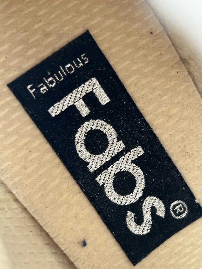 Puede incluir: Primer plano de una etiqueta rectangular negra con la palabra "Fabs" en letras grandes plateadas bordadas. Encima de "Fabs" est&aacute; la palabra "Fabulous" en letras plateadas bordadas m&aacute;s peque&ntilde;as. La etiqueta est&aacute; sobre una superficie beige.
