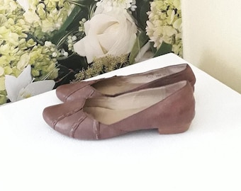 May include: A pair of brown leather flats with a low heel. The shoes have a rounded toe and decorative stitching across the vamp. The background features white and green floral arrangements.