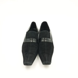Puede incluir: Un par de zapatos de ante negro con punta puntiaguda y una tira de cuero negro sobre el empeine.
