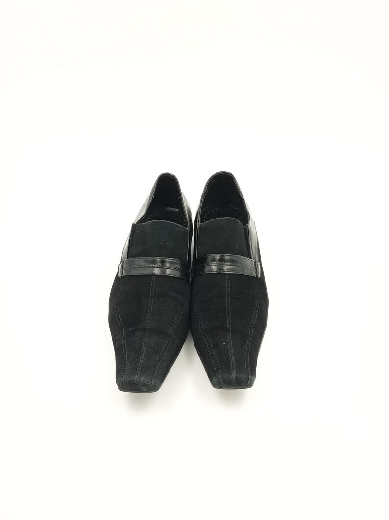 Puede incluir: Un par de zapatos de ante negro con punta puntiaguda y una tira de cuero negro sobre el empeine.