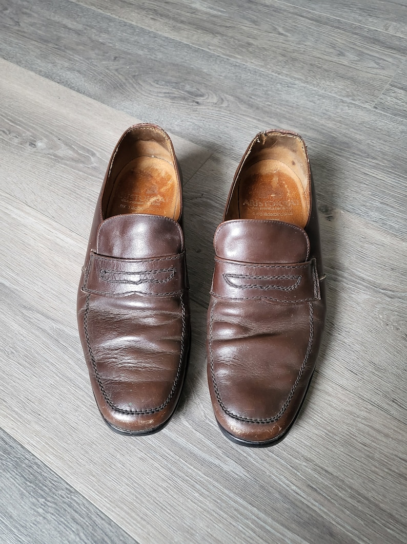 May include: A pair of brown leather loafers with a simple design. The loafers have a stitched seam around the edge of the shoe and a small strap across the top.